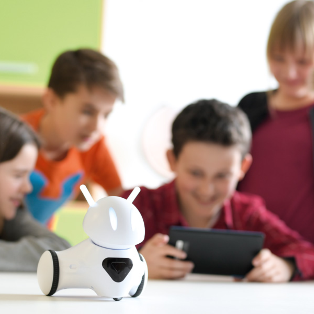 Dzieci programują robota Photon na tablecie. Z przodu biały robot ze świecącymi czułkami na stole, w tle rozmazane kontury dzieci.