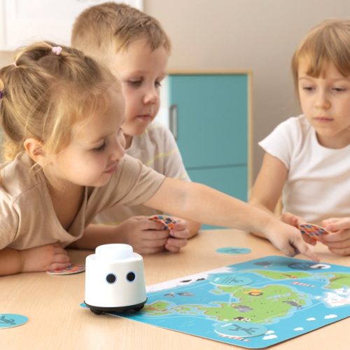 Dzieci siedzą przy stoliku i uczą się z robotem edukacyjnym Logi Mini Bot, korzystając z kolorowej maty edukacyjnej i kart do zabawy