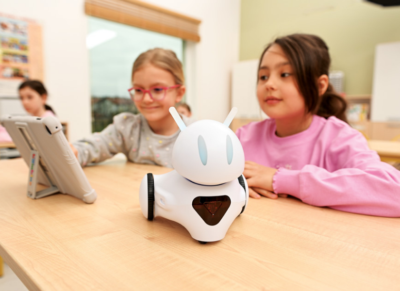 Dwie dziewczynki siedzą przy stoliku w sali lekcyjnej i obserwują robota edukacyjnego obok tabletu.